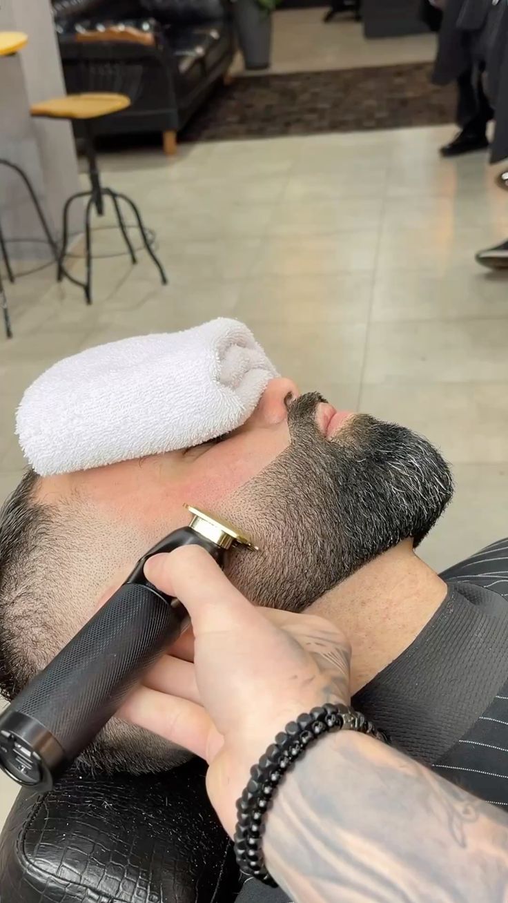 A barber detailing a beard with a trimmer and a hot towel